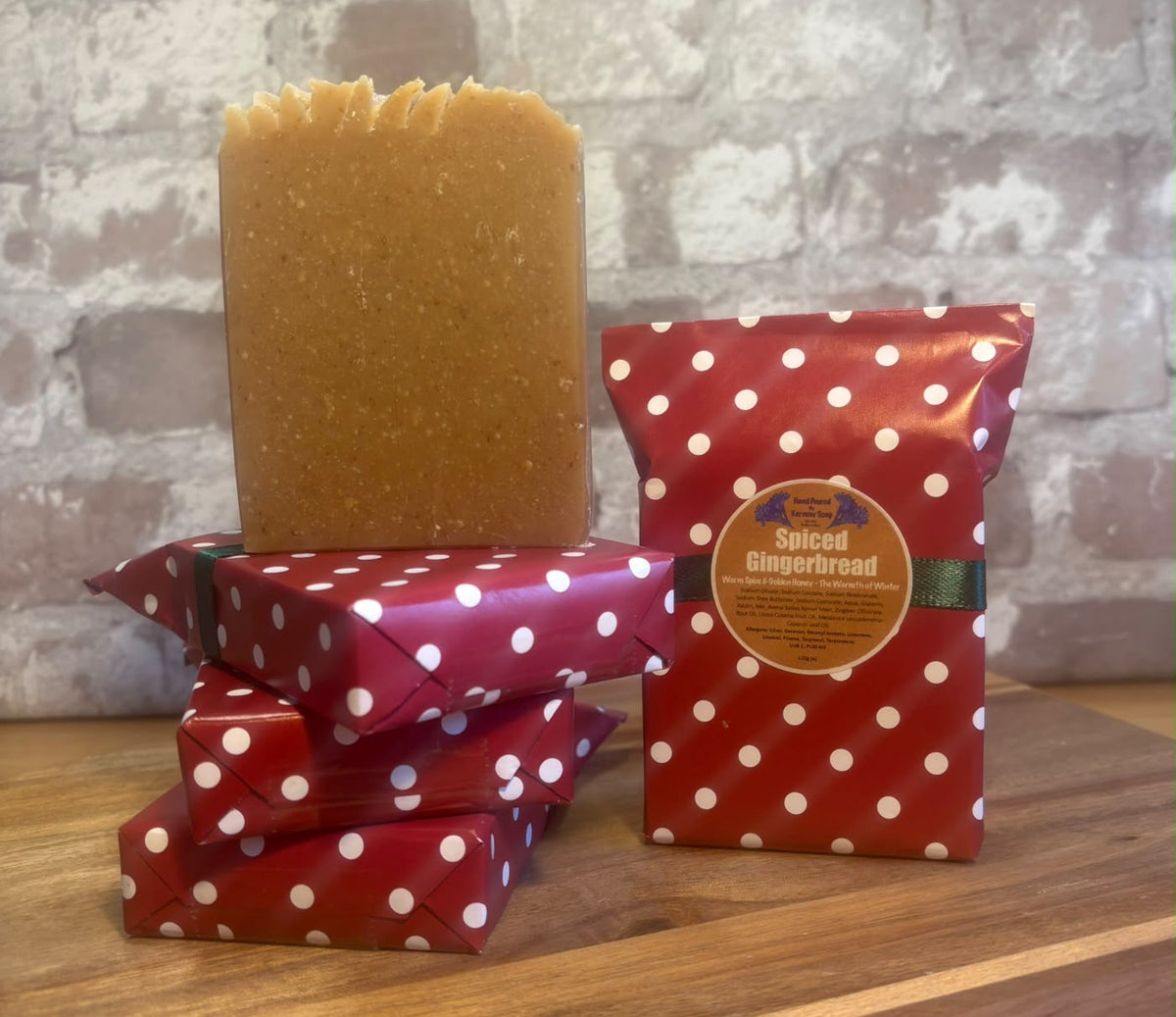 A bar of unwrapped gingerbread soap standing upright on a stack of three wrapped bars next to a bar  of 'Spiced Gingerbread' Kernow soap, all on a wooden surface against a rustic white brick wall.