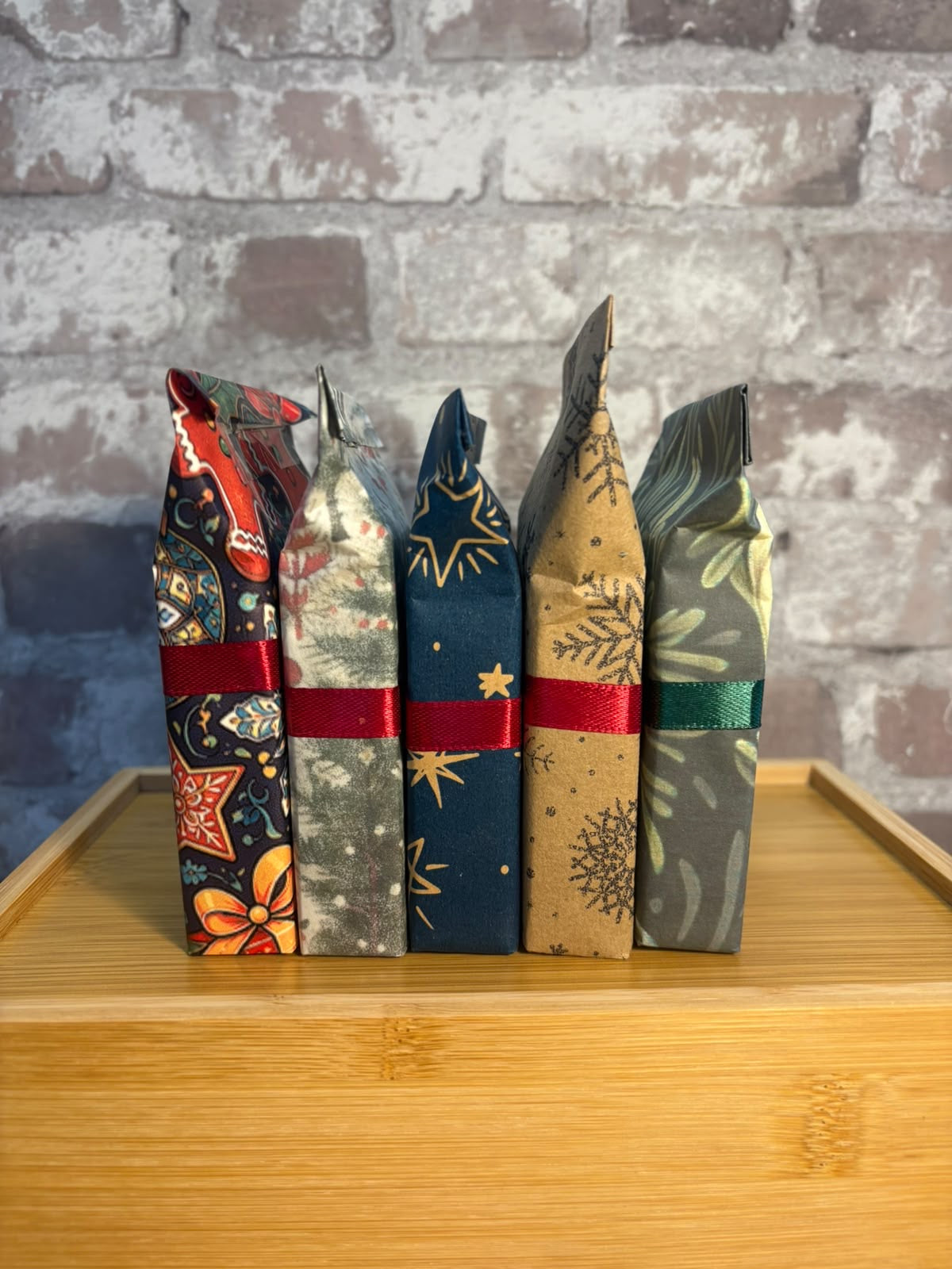 Five wrapped gift pouches or small packages standing upright on a wooden tray, each with a different holiday-themed pattern and tied with a red or green ribbon, set against a gray brick wall.