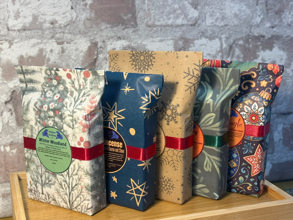 Five packaged soap bars standing upright on a wooden tray against a gray brick wall. The packages feature different holiday-themed patterns including snowflakes, stars, and floral designs, with red ribbons and circular labels.