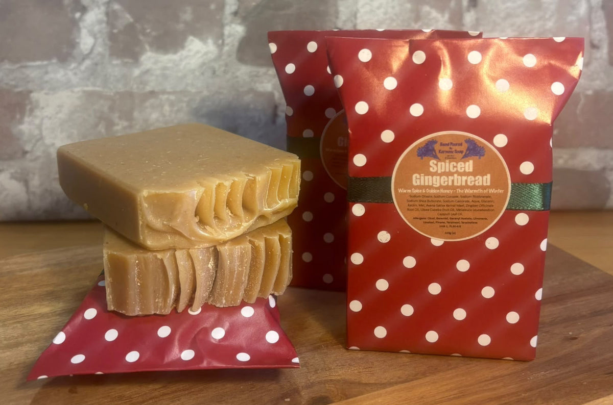 A stack of Spiced Gingerbread soap bars wrapped in red-and-white polka dot  Christmas themed paper  labeled 'Spiced Gingerbread' by Kernow Soap,  on a wooden surface against a faux brick wall background.