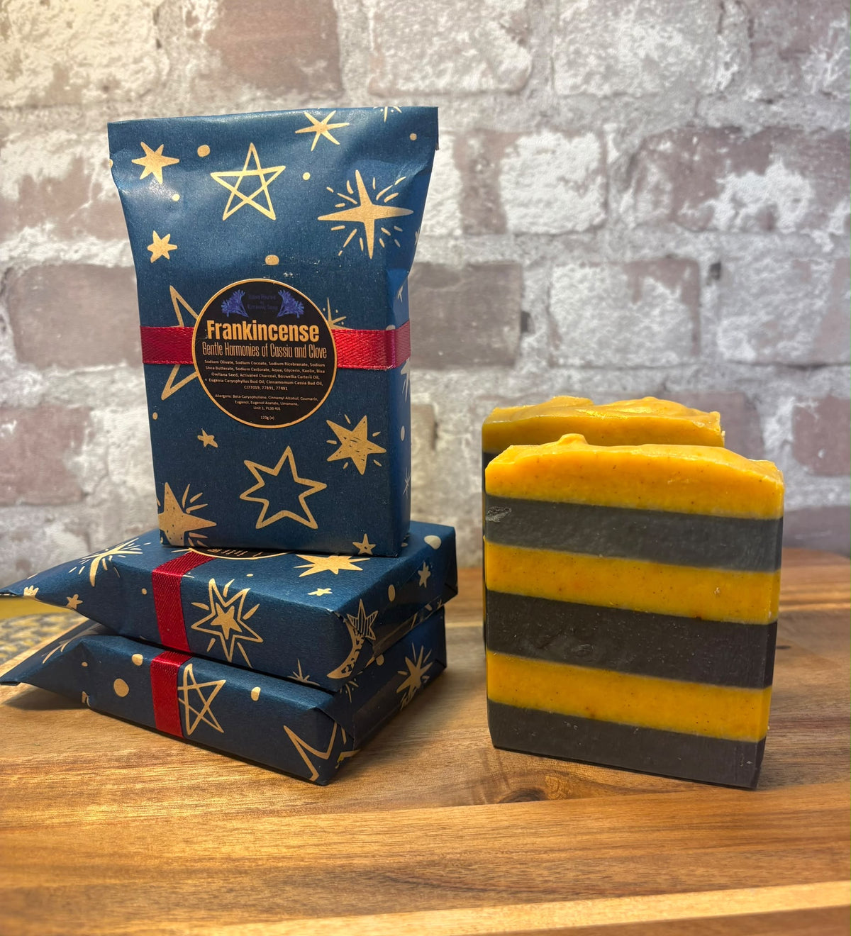 A packaged bar of Frankincense Kernow soap product shown on a wooden surface against a rustic brick wall. The set includes a tall blue gift bag with gold stars and a red ribbon, two matching wrapped bars, and two unwrapped rectangular soap bars with horizontal yellow and dark gray stripes.