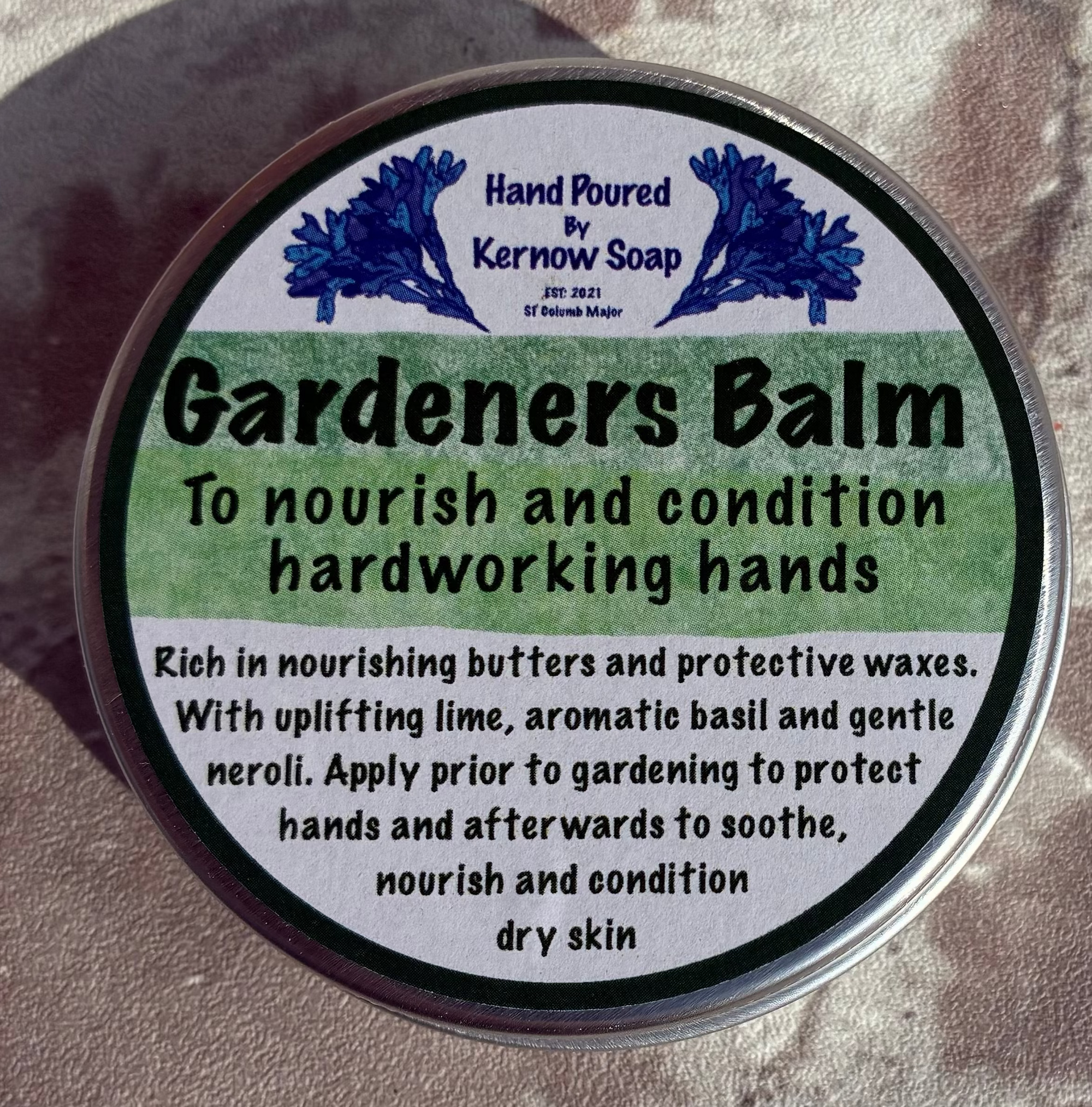 A round tin of Karmen Soap Gardeners Balm resting on a textured, weathered stone surface. The label is green and white with blue branding, and the tin has a silver metal rim.