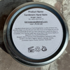 A round metal tin of Gardener's Hand Balm shown from the back, featuring a white label with product details, placed on a textured concrete surface.