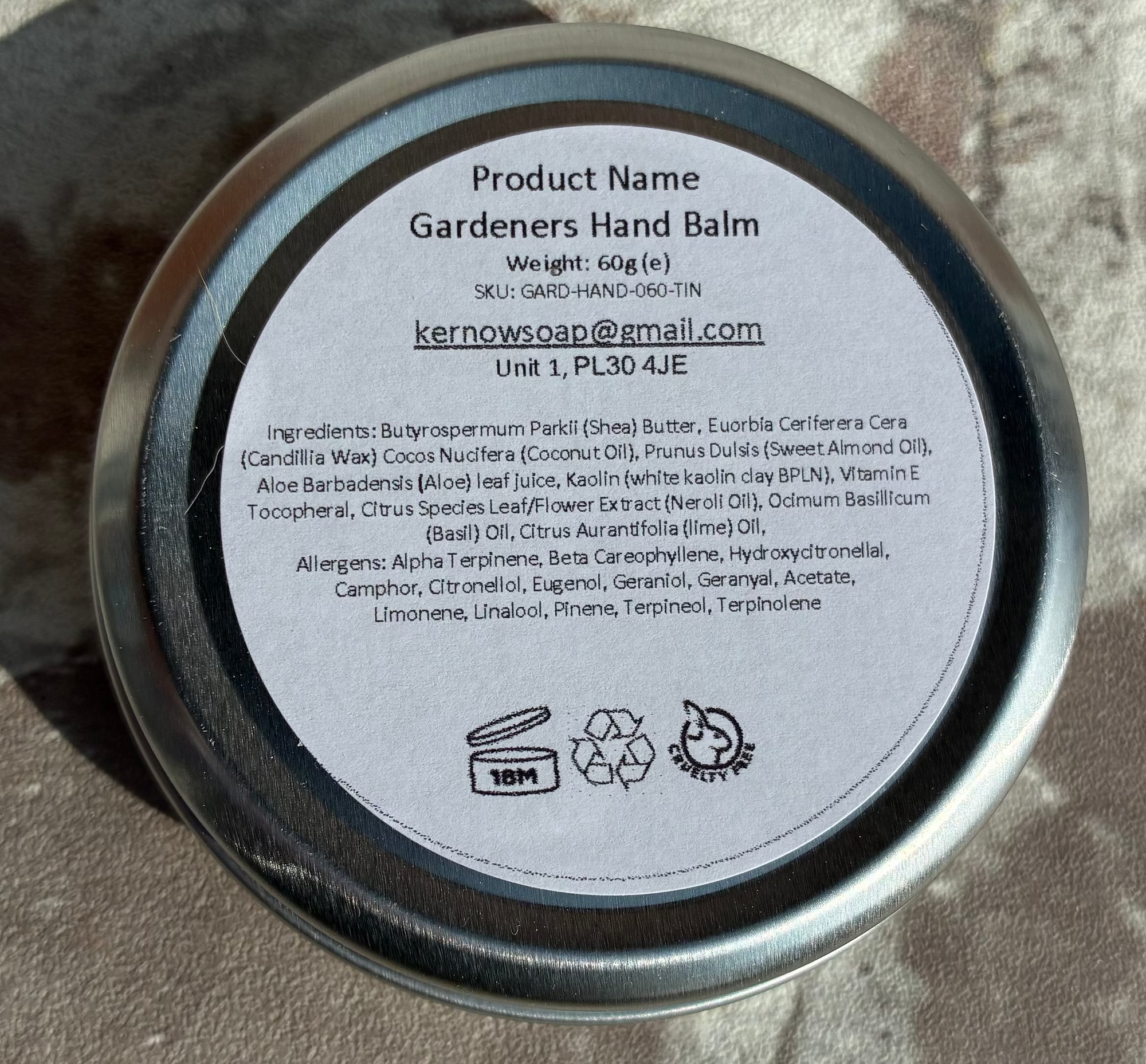A round metal tin of Gardener's Hand Balm shown from the back, featuring a white label with product details, placed on a textured concrete surface.