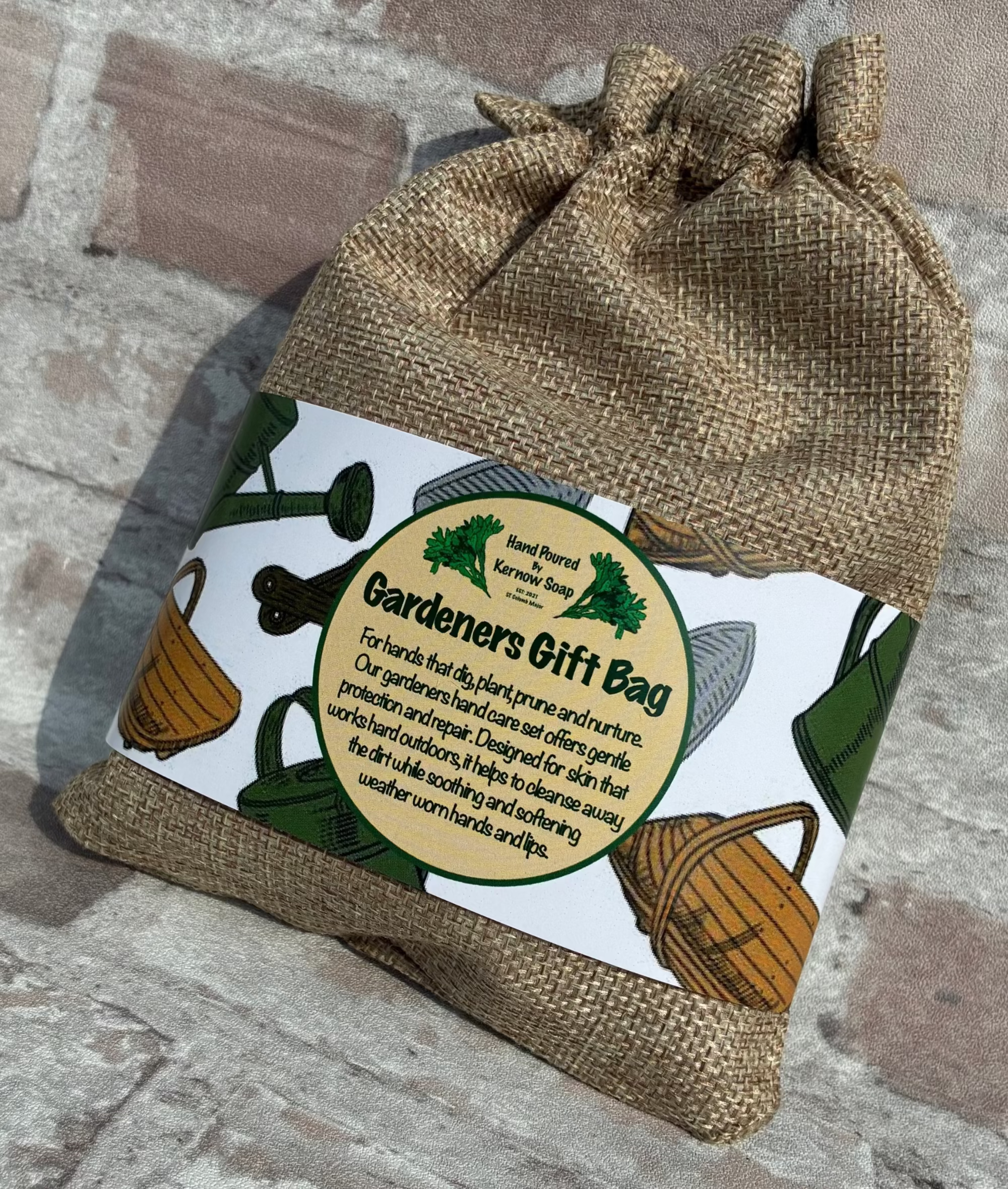 A burlap drawstring gift bag with a printed label reading 'Gardeners Gift Bag' and showing gardening illustrations, placed against a rustic brick wall.