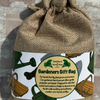 A burlap drawstring gift bag with a printed paper label reading 'Gardeners Gift Bag' and 'Hand Picked Farmhouse Gifts,' photographed against a weathered brick wall.