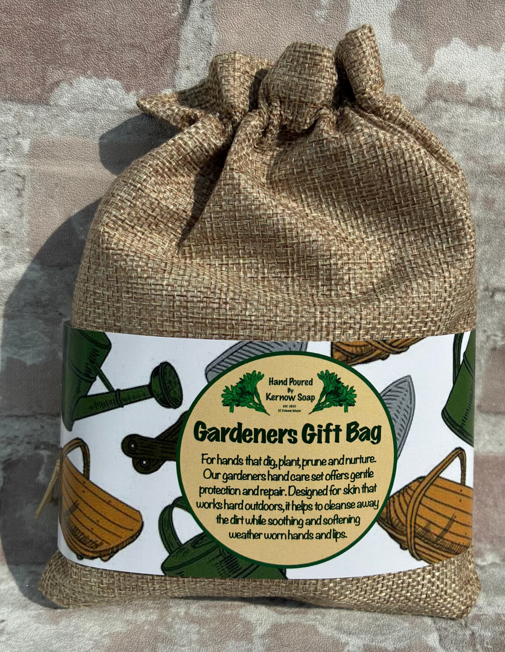 A burlap drawstring gift bag with a printed paper label reading 'Gardeners Gift Bag' and 'Hand Picked Farmhouse Gifts,' photographed against a weathered brick wall.