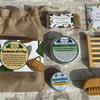A curated bath and body gift set arranged on a rustic stone surface, including a burlap drawstring pouch, a wooden soap brush, a wooden soap dish, two round soap tins, a small boxed item, and a floral card or booklet.