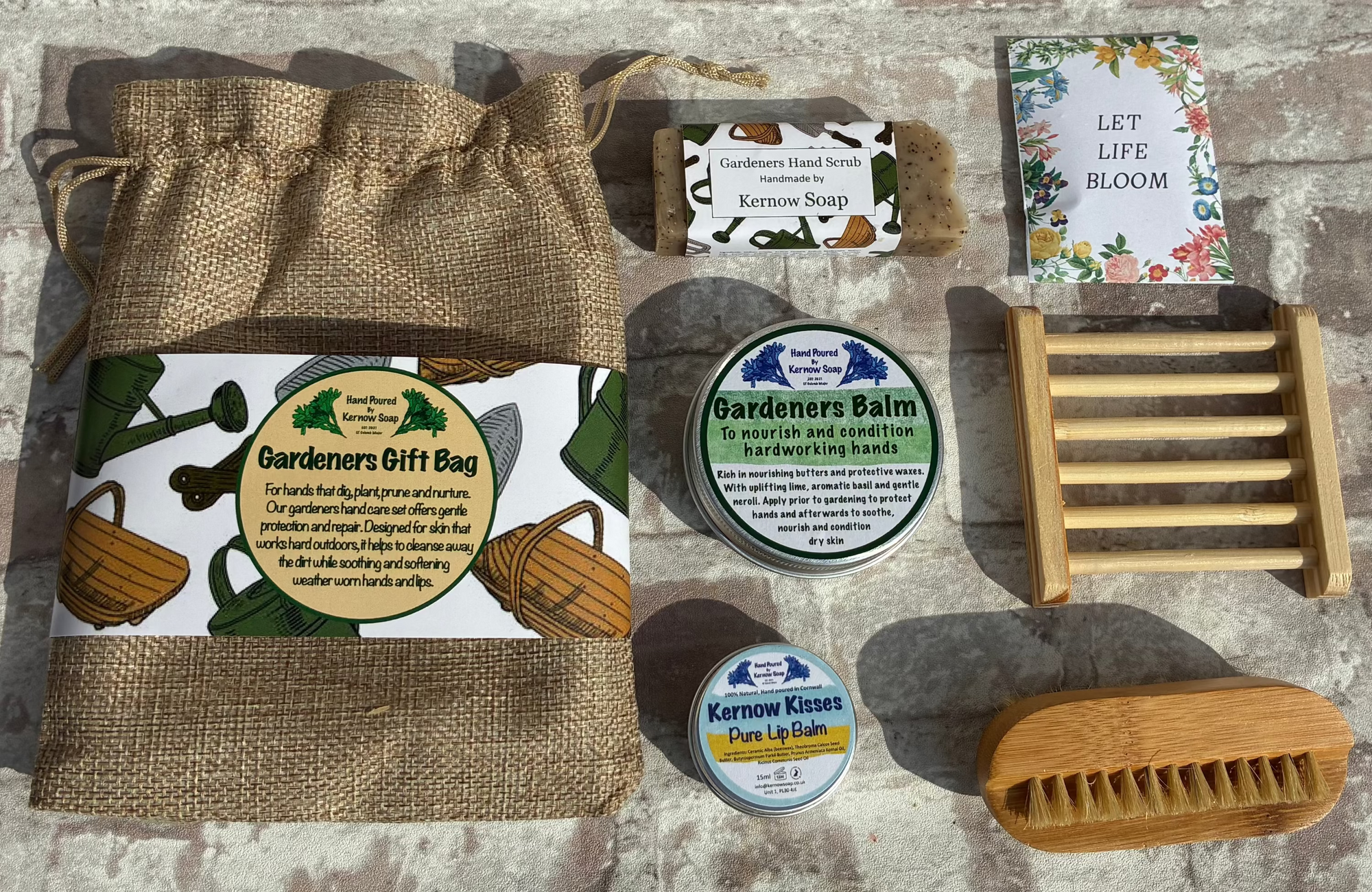 A curated bath and body gift set arranged on a rustic stone surface, including a burlap drawstring pouch, a wooden soap brush, a wooden soap dish, two round soap tins, a small boxed item, and a floral card or booklet.