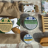 A curated gift set arranged on a rustic stone surface, including a burlap drawstring bag with a botanical label, two small round tins, a wooden scrubber, a wooden soap dish, and a floral card or booklet.