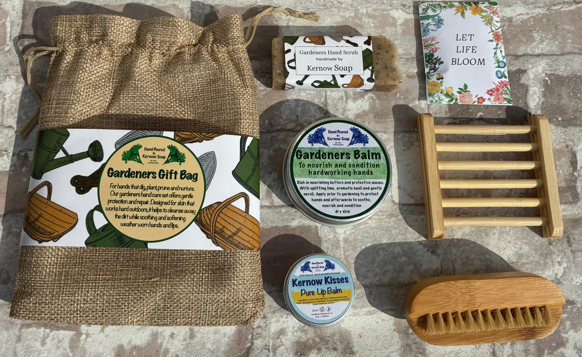 A curated gift set arranged on a rustic stone surface, including a burlap drawstring bag with a botanical label, two small round tins, a wooden scrubber, a wooden soap dish, and a floral card or booklet.