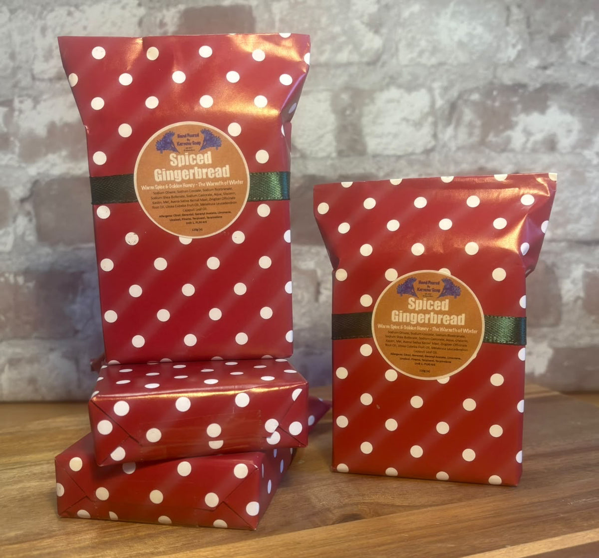 Three bars of Spiced Gingerbread soap from Kernow Soap, wrapped in red and white polka dot paper with green ribbons and round brown labels, displayed on a wooden surface against a rustic brick wall.