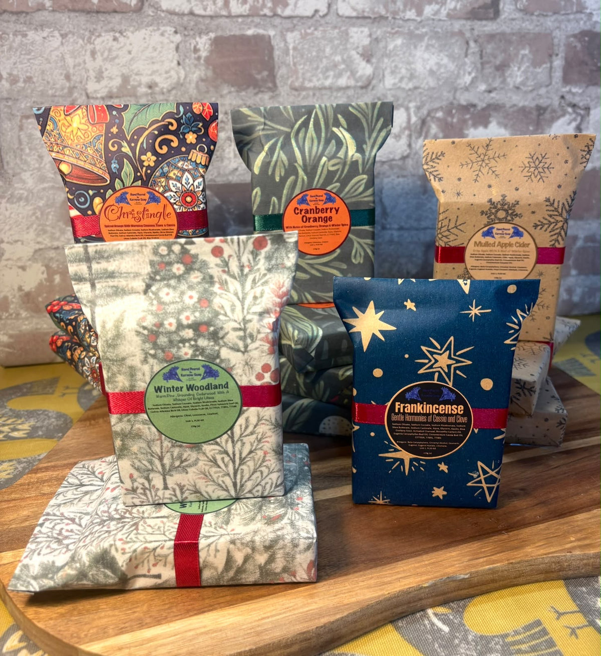 Six packaged Christmas themed soap bars from Kernow Soap are arranged on a wooden board in front of a brick wall. The packages feature festive Christmas patterns including snowflakes, holly, pine trees, and paisley, with labels showing the tea names: Christingle, Cranberry Orange, Winter Wonderland, and frankincense.