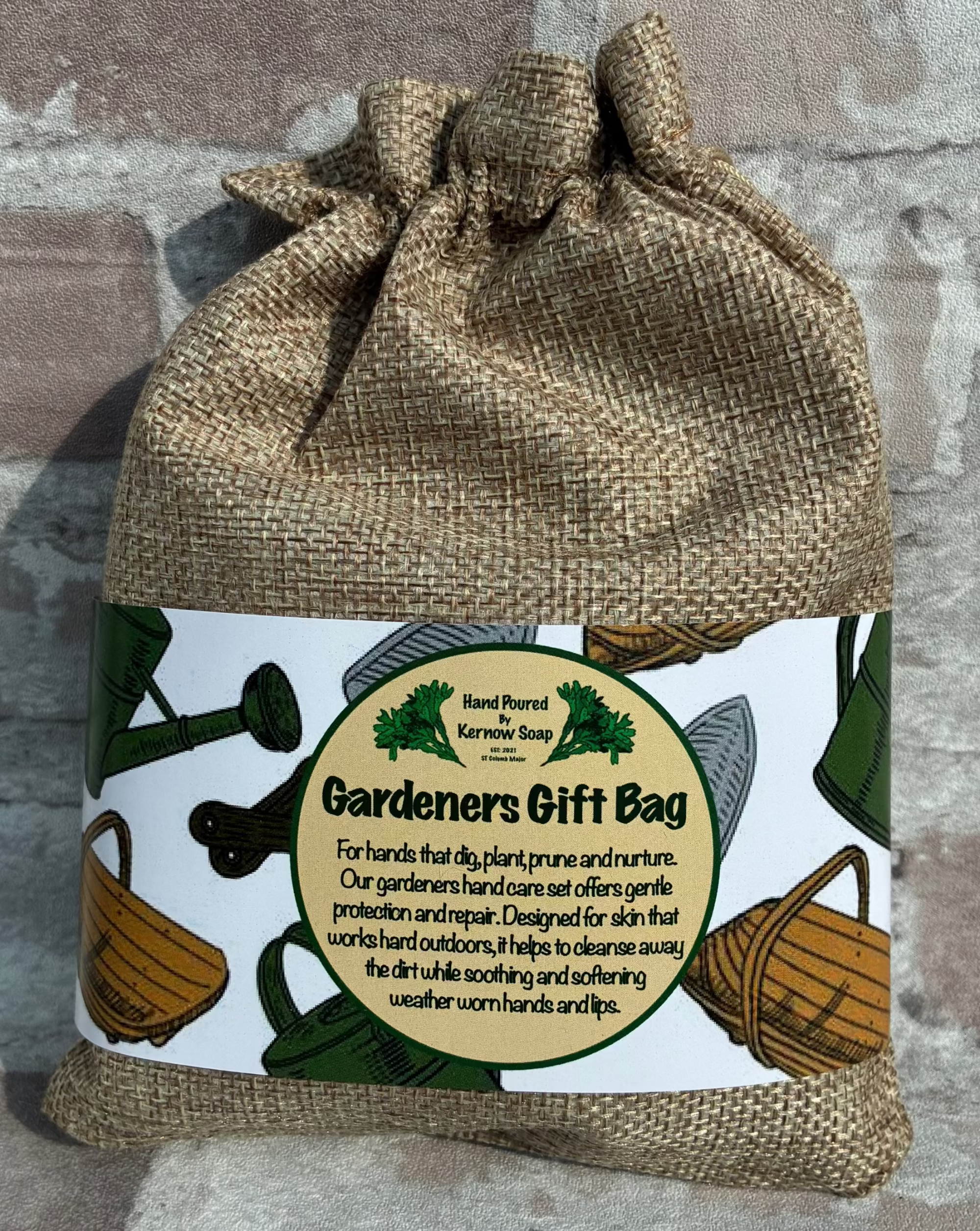 A burlap drawstring gift bag with a printed paper label reading 'Gardeners Gift Bag' and 'Hand Poured by Kernow Soap,' photographed against a stone wall background.