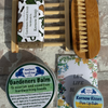 A flat-lay of a hand and face care set on a gray stone-textured background, featuring a bamboo hand brush, a wooden soap holder with a bar of soap labeled Kermow Soap, two tins of Kermow Kisses Pure Lip Balm, and a bar of Gardeners Balm.