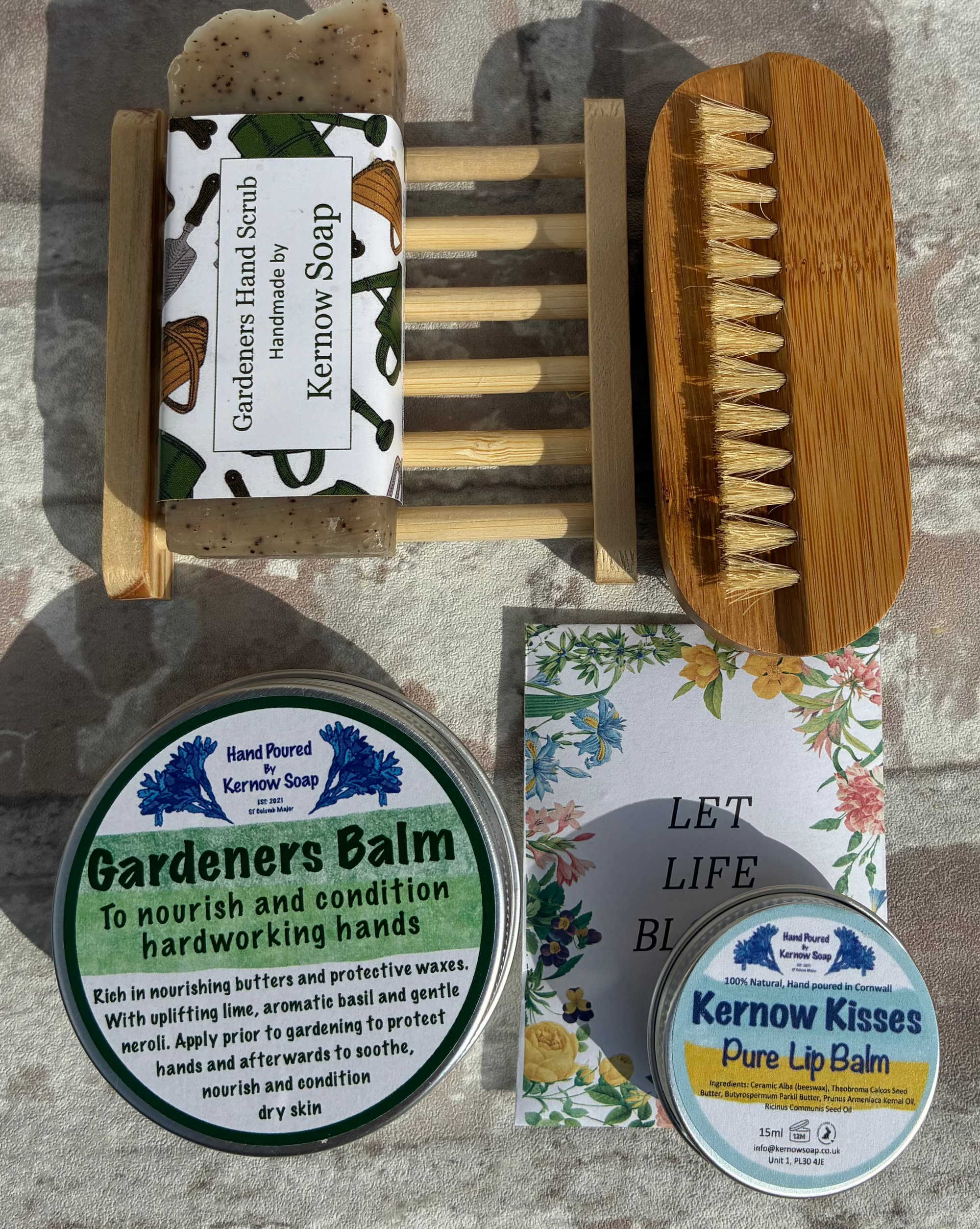 A flat-lay of a hand and face care set on a gray stone-textured background, featuring a bamboo hand brush, a wooden soap holder with a bar of soap labeled Kermow Soap, two tins of Kermow Kisses Pure Lip Balm, and a bar of Gardeners Balm.