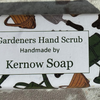 A bar of handmade gardeners' hand scrub soap with a white paper label, placed on a textured stone surface.