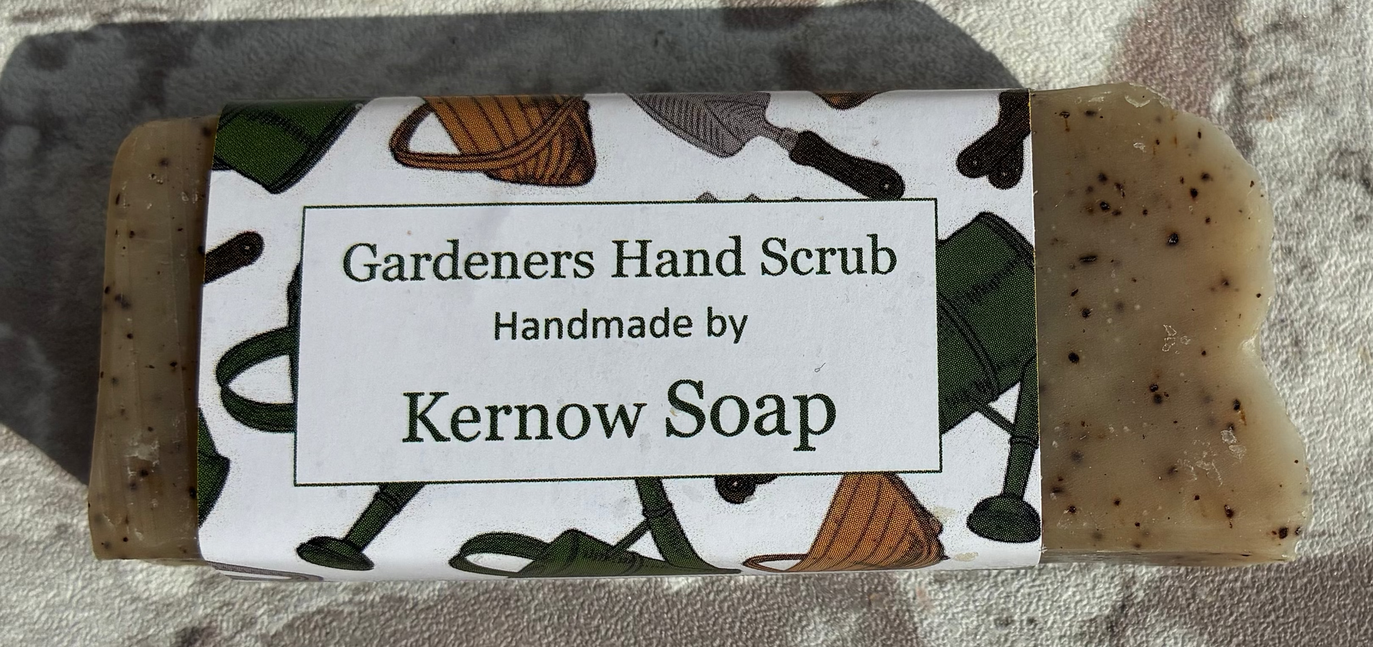 A bar of handmade gardeners' hand scrub soap with a white paper label, placed on a textured stone surface.
