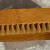 A natural bamboo-handled brush with short tan bristles, photographed from above on a textured light gray surface.