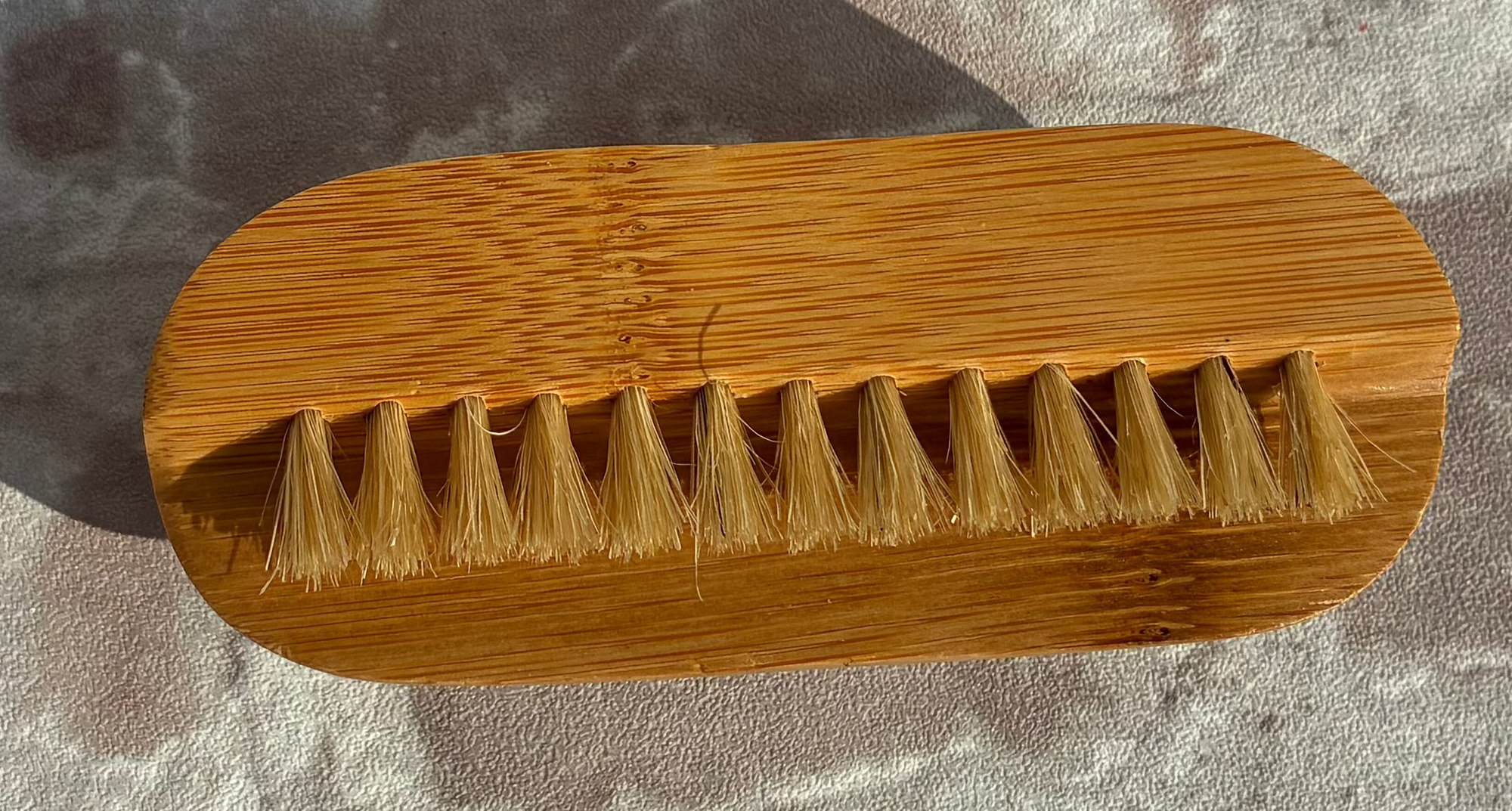 A natural bamboo-handled brush with short tan bristles, photographed from above on a textured light gray surface.