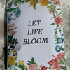 A rectangular printed card or notebook cover with the words “LET LIFE BLOOM” in black serif text, surrounded by a colorful floral border, placed on a textured stone surface.