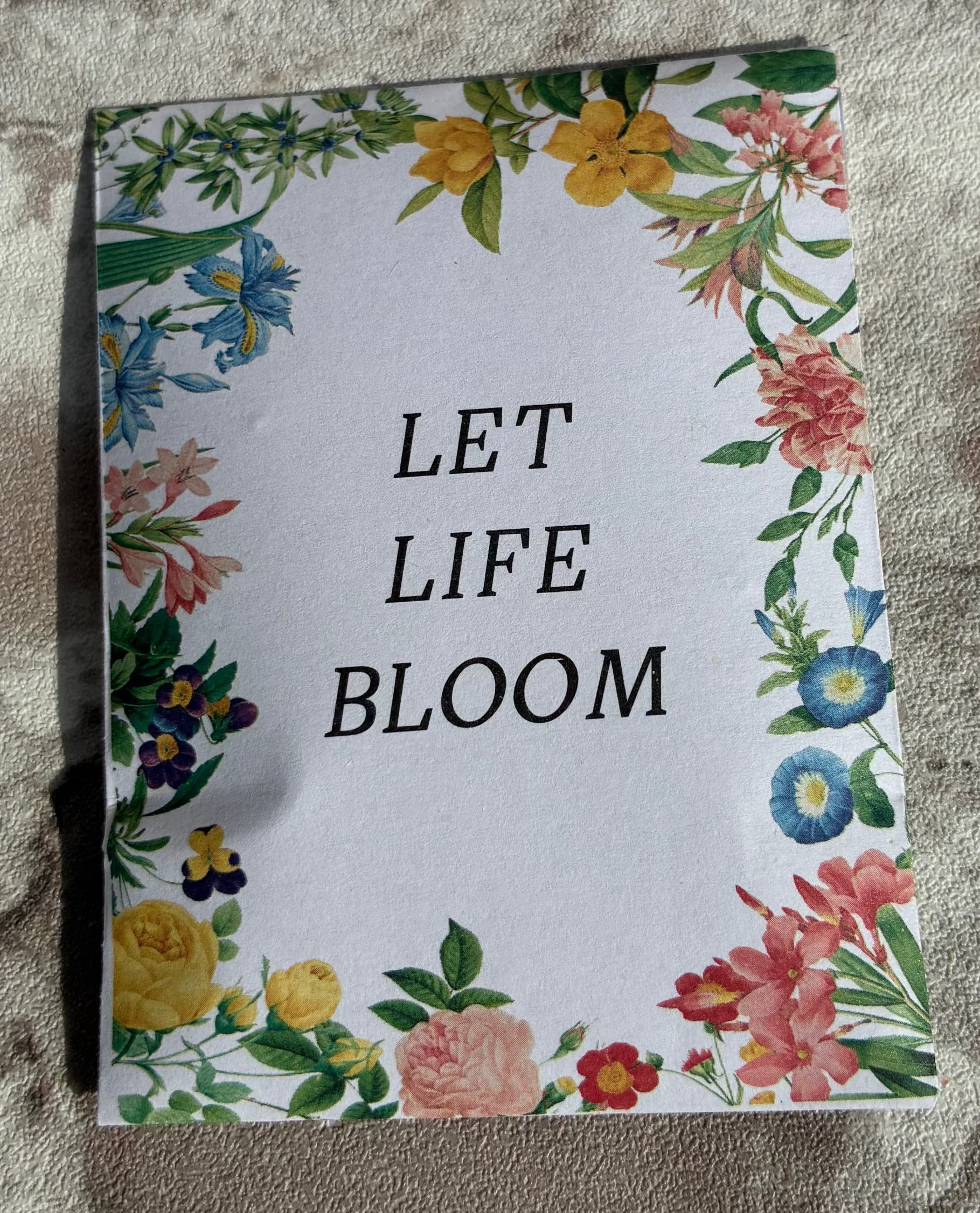 A rectangular printed card or notebook cover with the words “LET LIFE BLOOM” in black serif text, surrounded by a colorful floral border, placed on a textured stone surface.
