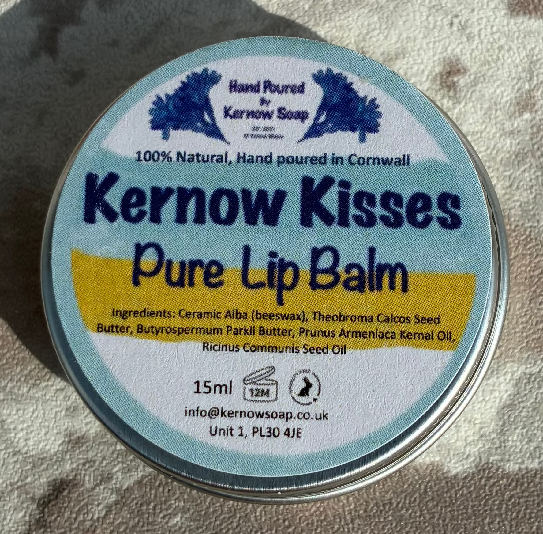 A round tin of Kernow Kisses Pure Lip Balm resting on a textured, light-colored brick surface. The tin features a white label with blue text and a yellow accent stripe.
