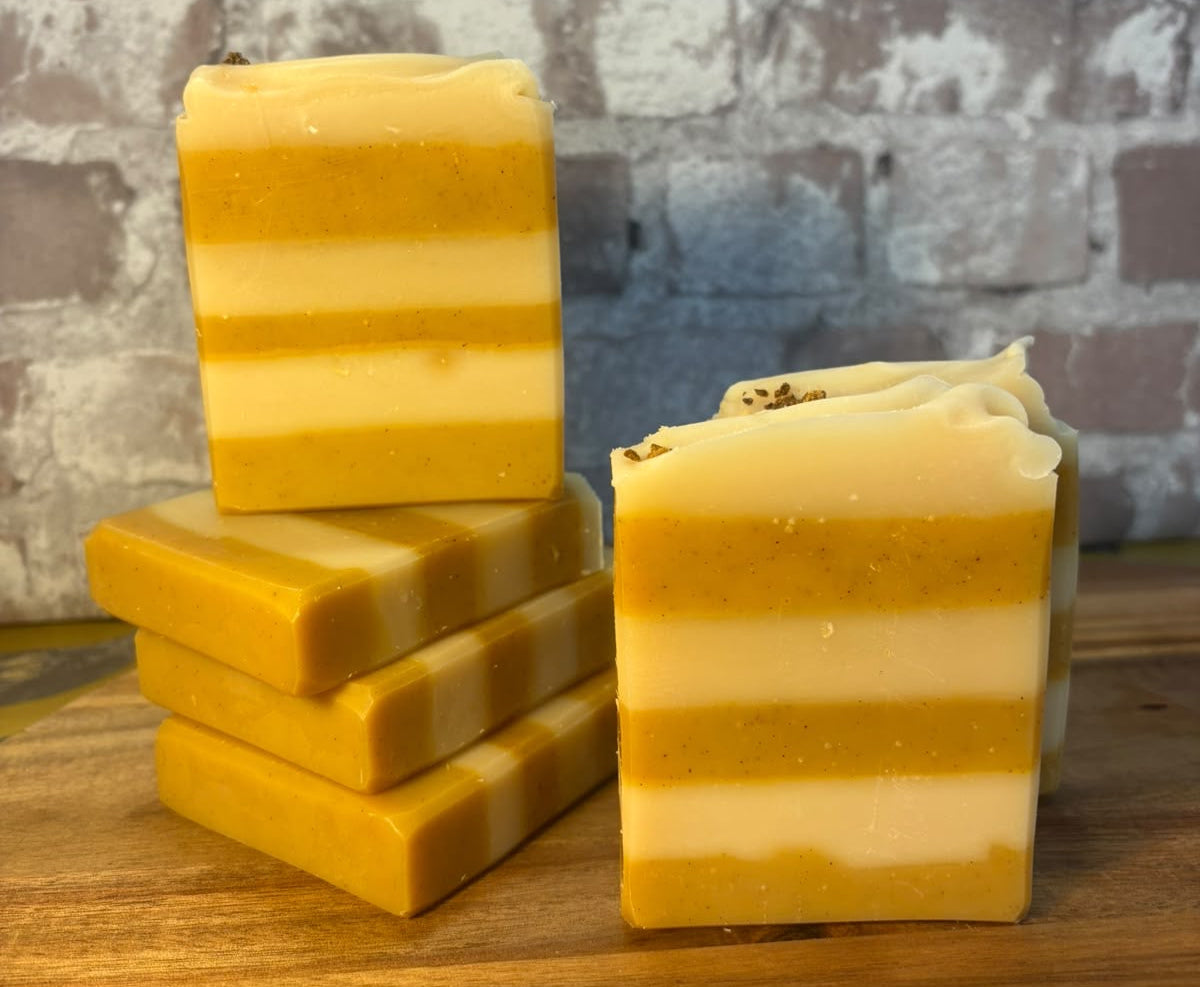 Three handmade striped soap bars in yellow and cream tones, with two stacked on the left and one standing on the right, displayed on a wooden surface against a rustic brick wall background.